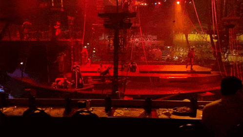 a man sits at a table in front of a stage with a red light behind him