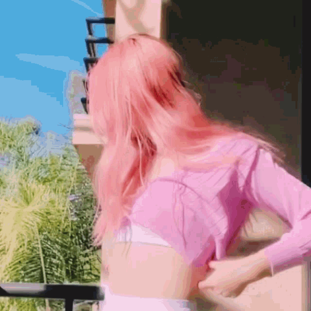a woman with pink hair is standing on a balcony wearing a pink top and a white bra .