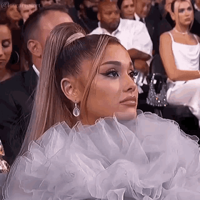 ariana grande is wearing a white dress and earrings while sitting in the audience at awards .