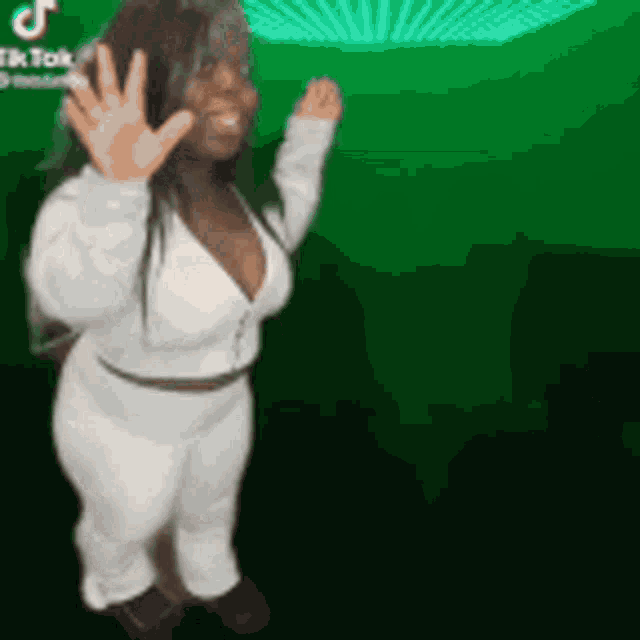 a woman in a white outfit is standing in front of a green background and waving her hand .