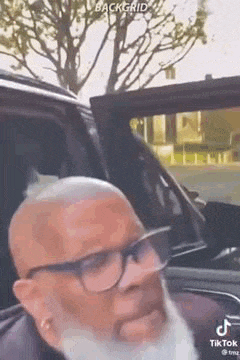 a bald man with glasses and a beard is sitting in the back seat of a car .