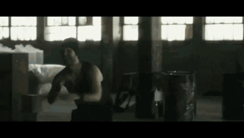 a man in a black tank top is squatting in a dark room with a pepsi barrel in the background