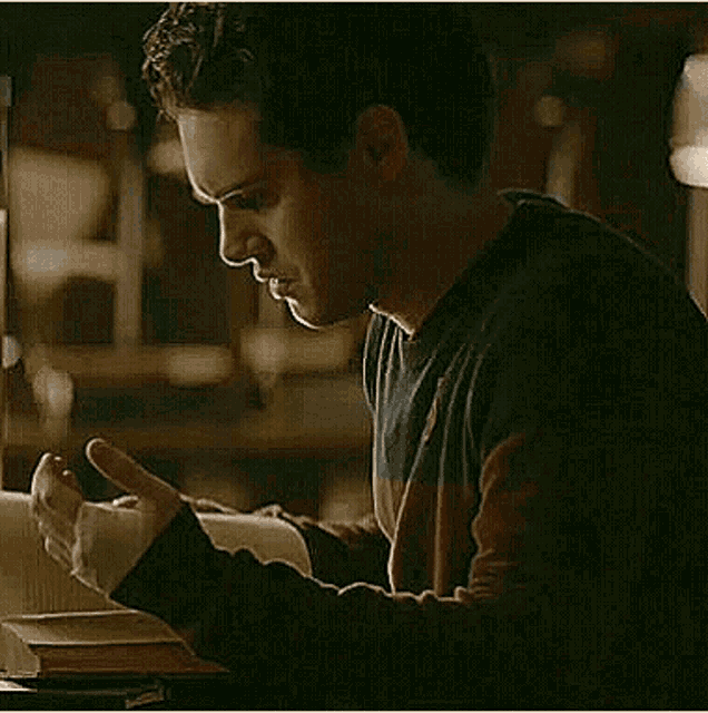 a man is reading a book while sitting at a table in a dark room .