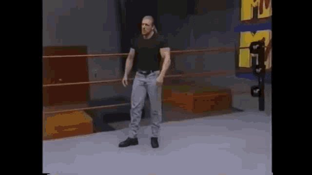 a man is standing in a wrestling ring wearing jeans and a black shirt .