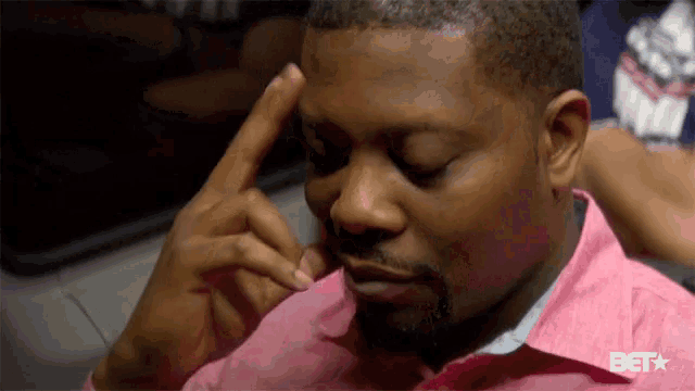 a man in a pink shirt is scratching his forehead with his finger .