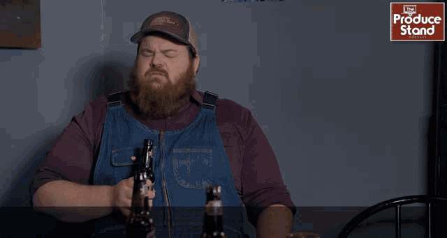 a man with a beard is sitting at a table with a bottle of beer and a sign that says produce stand