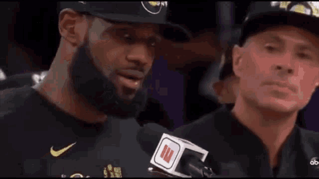 a man with a beard is talking into a microphone while another man watches .