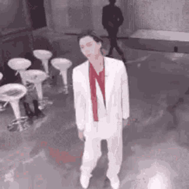 a man in a white suit and red tie is dancing in a room with stools .