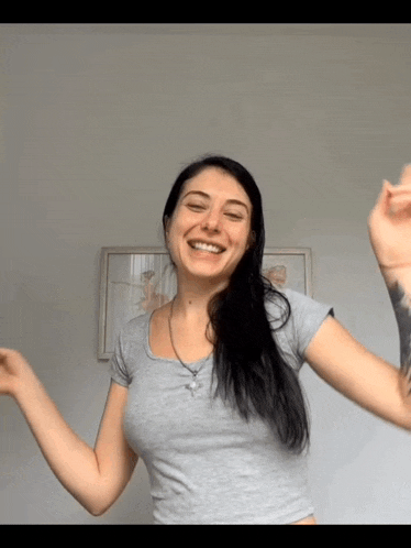 a woman in a grey shirt is smiling with her arms outstretched in front of a picture on the wall .
