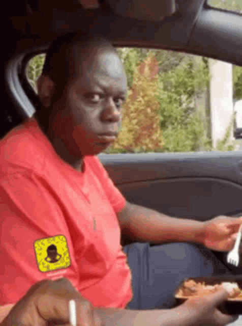 a man in a red shirt with a snapchat logo on his sleeve is sitting in a car eating food
