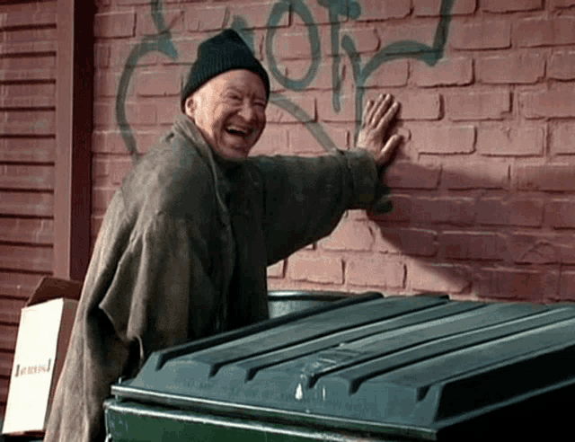 a man laughs in front of a brick wall that has graffiti on it that says " more "