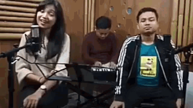 a man and a woman are singing into microphones in a recording studio while a man plays a keyboard .