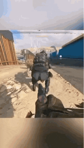 a soldier is running down a dirt road in a video game while another soldier watches .