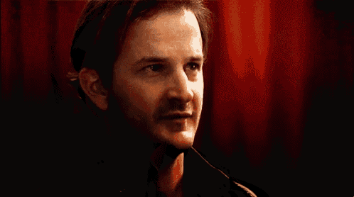 a man with a mustache is sitting in front of a red curtain .