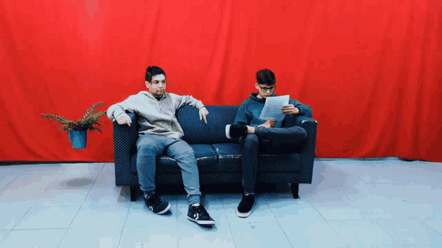 two young men are sitting on a couch reading papers