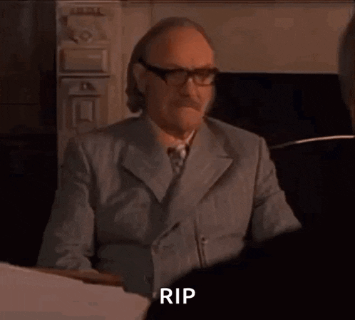 a man in a suit and tie is sitting in front of a table and says rip .