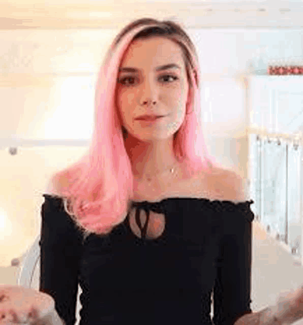 a woman with pink hair wearing a black off the shoulder top is standing in a room .