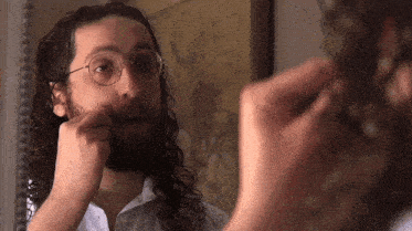 a man with long curly hair and glasses looks at himself in a mirror
