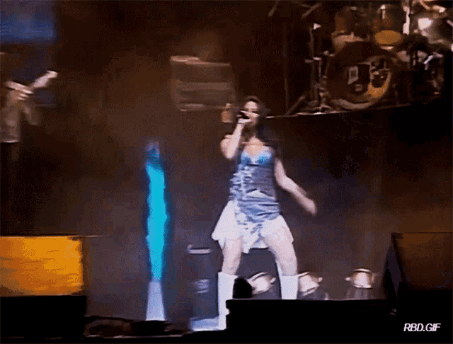 a woman singing into a microphone on a stage with a drum set behind her