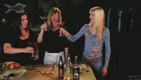 three women toasting with drinks in front of a sign that says " hungry "