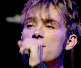 a close up of a man singing into a microphone with purple lights behind him