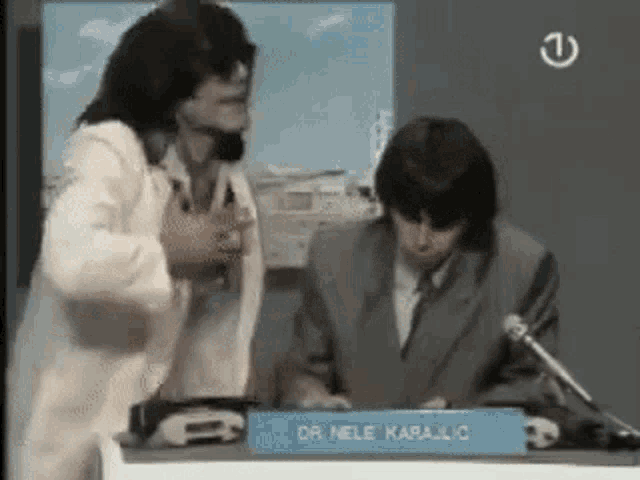 a man in a suit is sitting at a table with a sign that says dr. nele karajlic