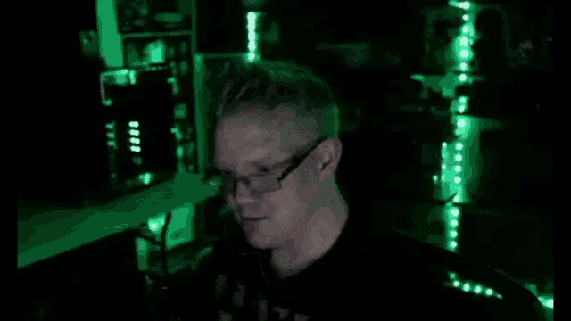 a man wearing glasses is laughing in a dark room with green lights behind him .