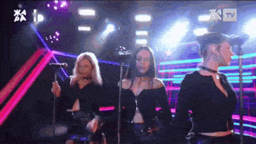 a group of women singing on a stage with a tv logo in the background