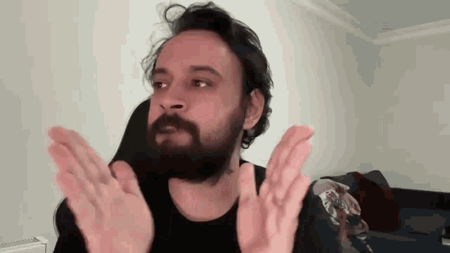 a man with a beard is sitting in a living room with his hands up .