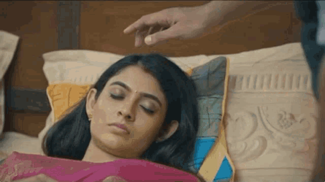 a woman is laying on a bed with her eyes closed and a man 's hand on her head .