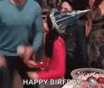 a woman is sitting at a table with a birthday hat on her head and a group of people are standing around her .