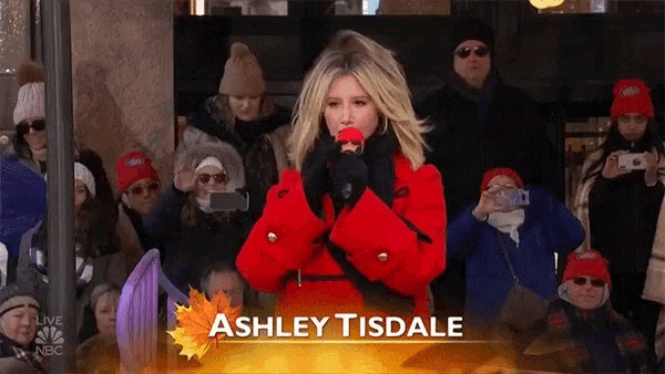 a woman in a red coat is standing in front of a crowd and says ashley tisdale