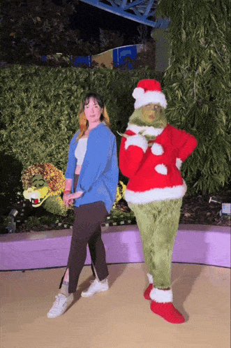 a woman stands next to a grinch mascot in a santa hat