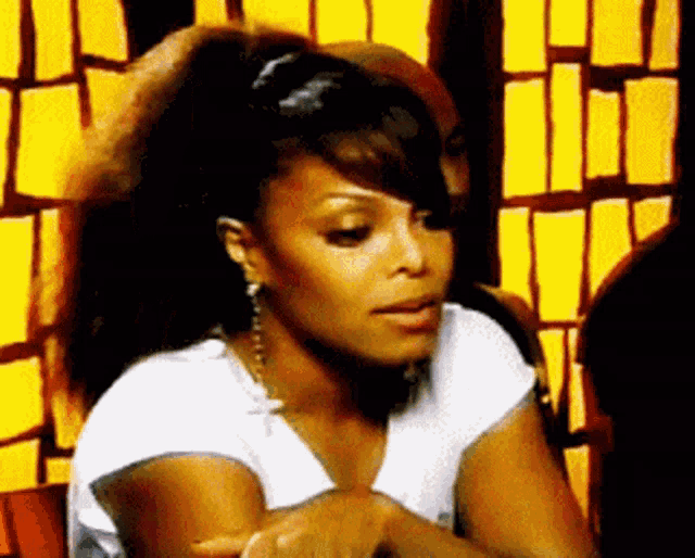 a woman in a white shirt is sitting in front of a yellow wall with a man in the background .