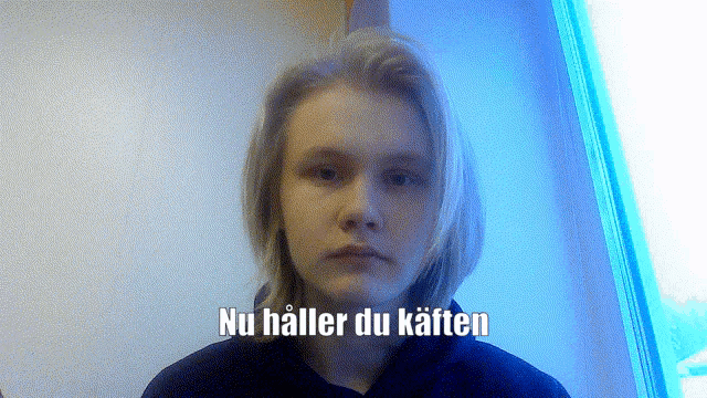 a young man with long blonde hair is standing in front of a window with the words nu håller du kaften above him