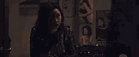 a woman is sitting at a table in a dark room with posters on the wall .