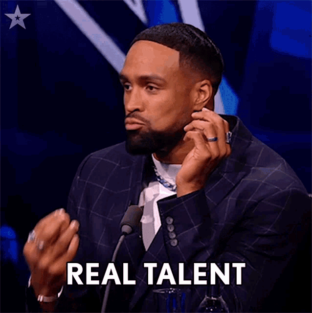 a man in a suit is speaking into a microphone and the word real talent is on the screen behind him
