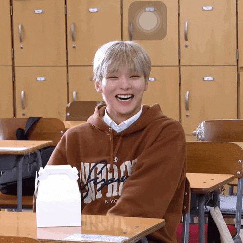 a boy wearing a brown nyc hoodie sits at a desk in a classroom