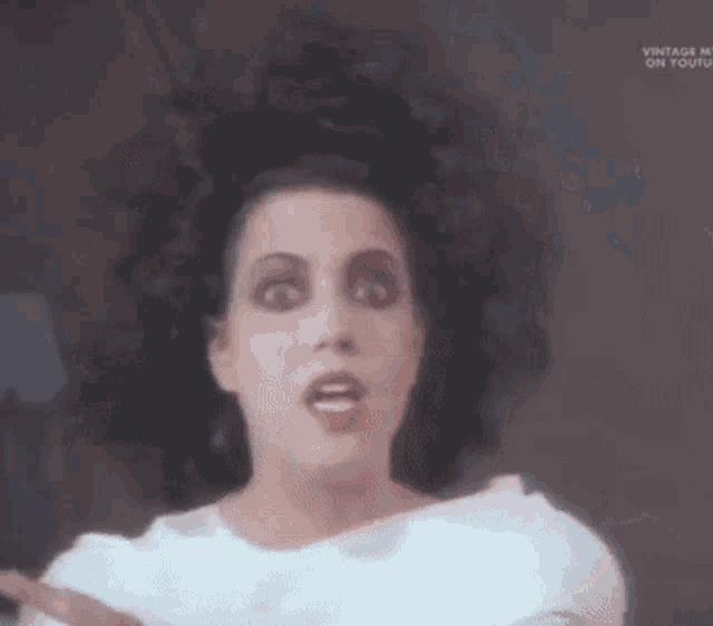 a woman with black hair and red lips is wearing a white shirt and making a surprised face .