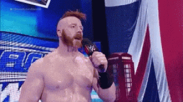 a shirtless wrestler with a beard is holding a microphone in front of a british flag .