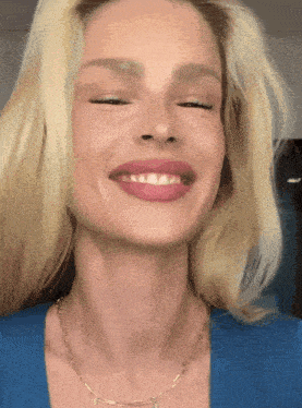 a blonde woman wearing a blue shirt and a gold chain smiles