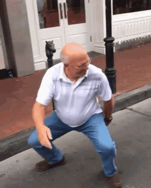 an older man in a white shirt and blue jeans is squatting down on the sidewalk