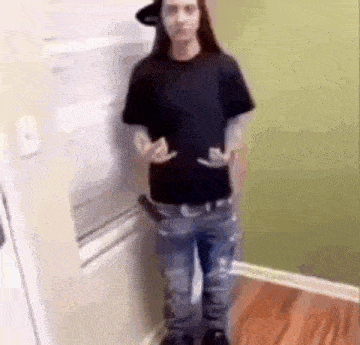 a man in a black shirt and blue jeans is standing in a kitchen holding a gun .