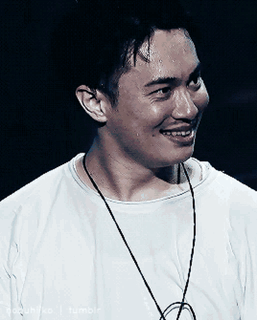 a man wearing a white shirt and a black necklace smiles for the camera