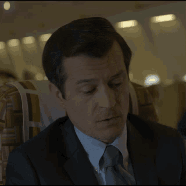 a man in a suit and tie sits on an airplane covering his mouth