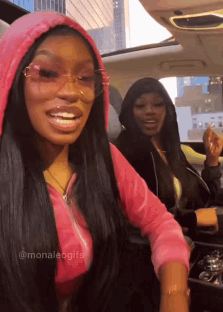a woman wearing a pink hoodie and sunglasses is sitting in a car next to another woman