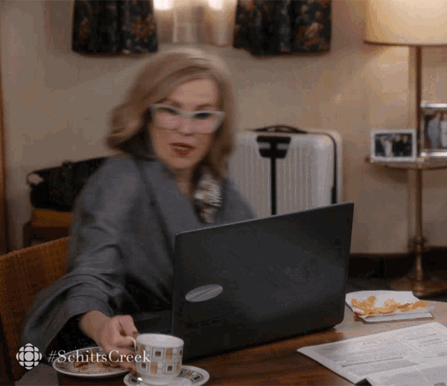 a woman sits at a table with a laptop and a cup of coffee with the hashtag schitts creek on the bottom