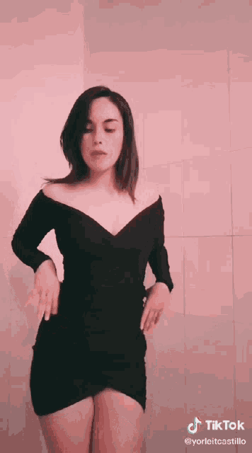 a woman in a short black dress is dancing in front of a pink wall with tiktok written on the bottom