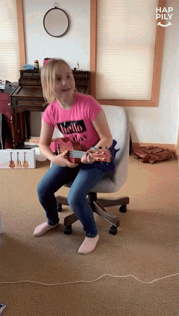 a girl in a pink shirt with the word hello on it sits in an office chair playing a guitar