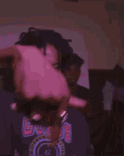 a young man with dreadlocks is wearing a purple shirt and making a funny face with his hand .